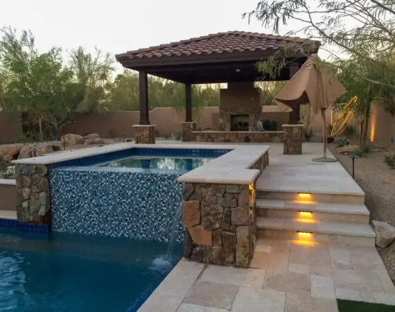 Outdoor spa with stone detailing and waterfall, illuminated steps leading to a shaded pavilion with a fireplace.