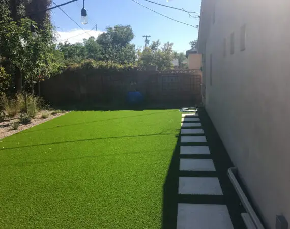 Backyard with artificial turf and a walkway of square stone pavers, lined with string lights.