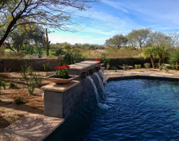 Scenic backyard water feature with multiple streams cascading into a pool, surrounded by red flowers, desert plants, and a cactus backdrop.