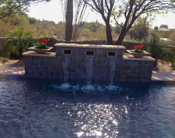 Decorative tile water feature with three flowing streams into a pool, flanked by red flowers and desert plants in a serene desert setting.