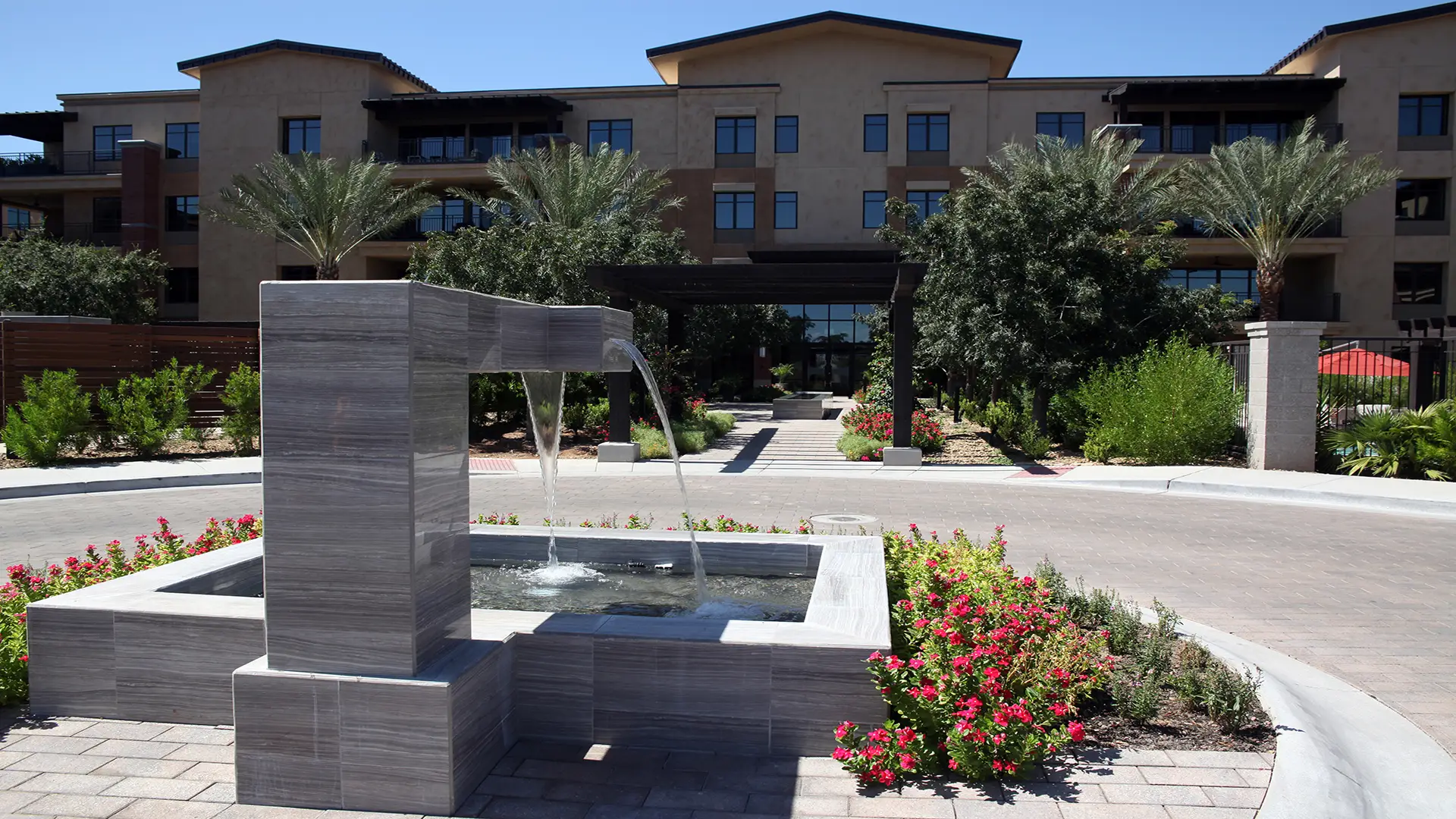 Modern apartment building entrance with a sleek water fountain, lush landscaping, and a pathway leading to the main entrance.