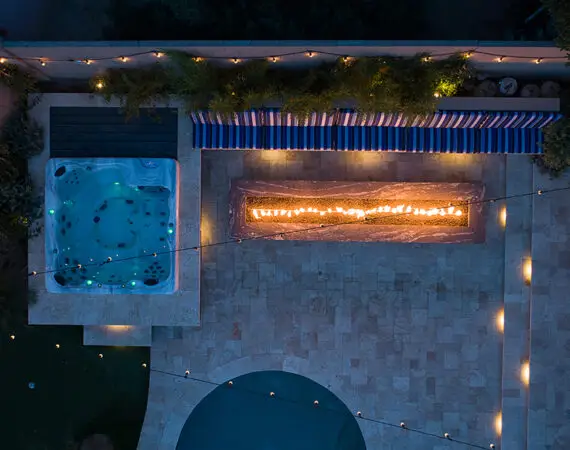 Aerial night view of a backyard featuring a lit fire feature, illuminated spa, and patio area with string lights.