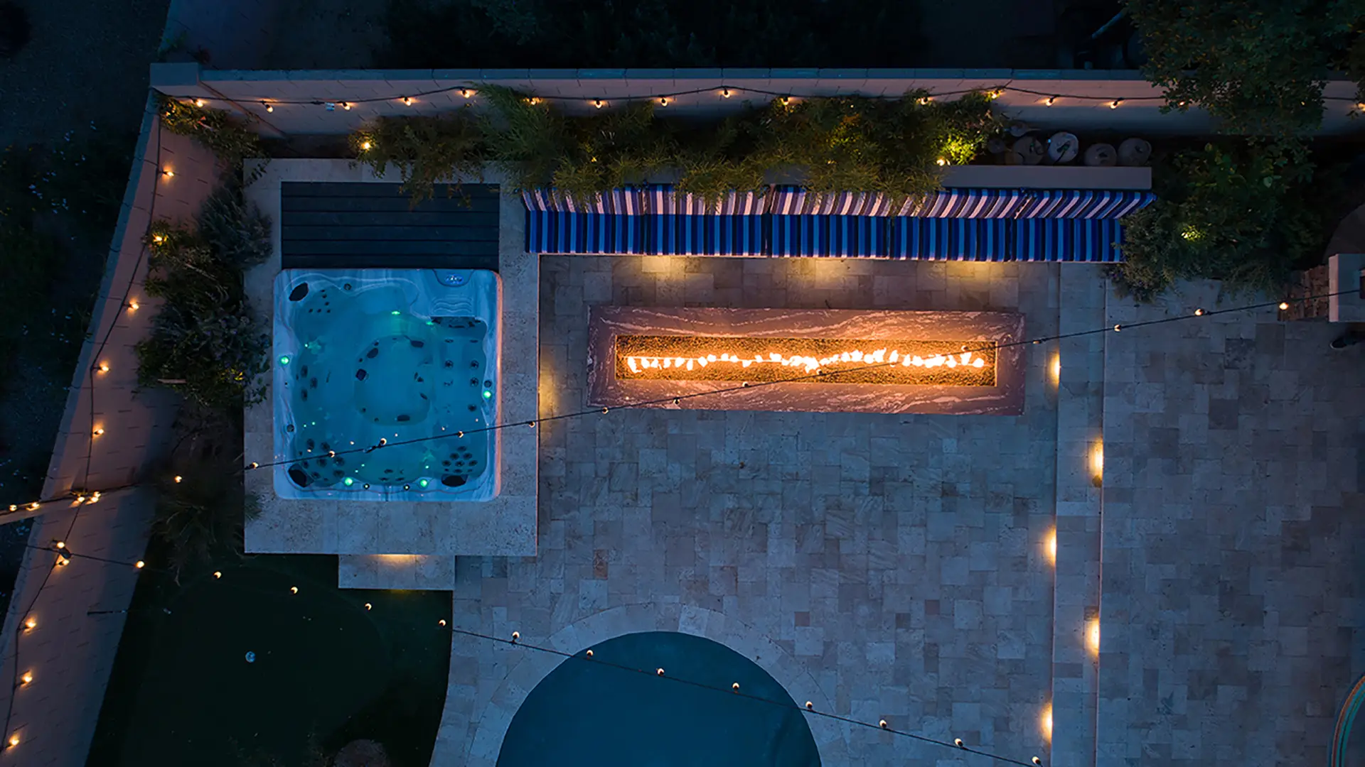 Aerial night view of a backyard featuring a lit fire feature, illuminated spa, and patio area with string lights.