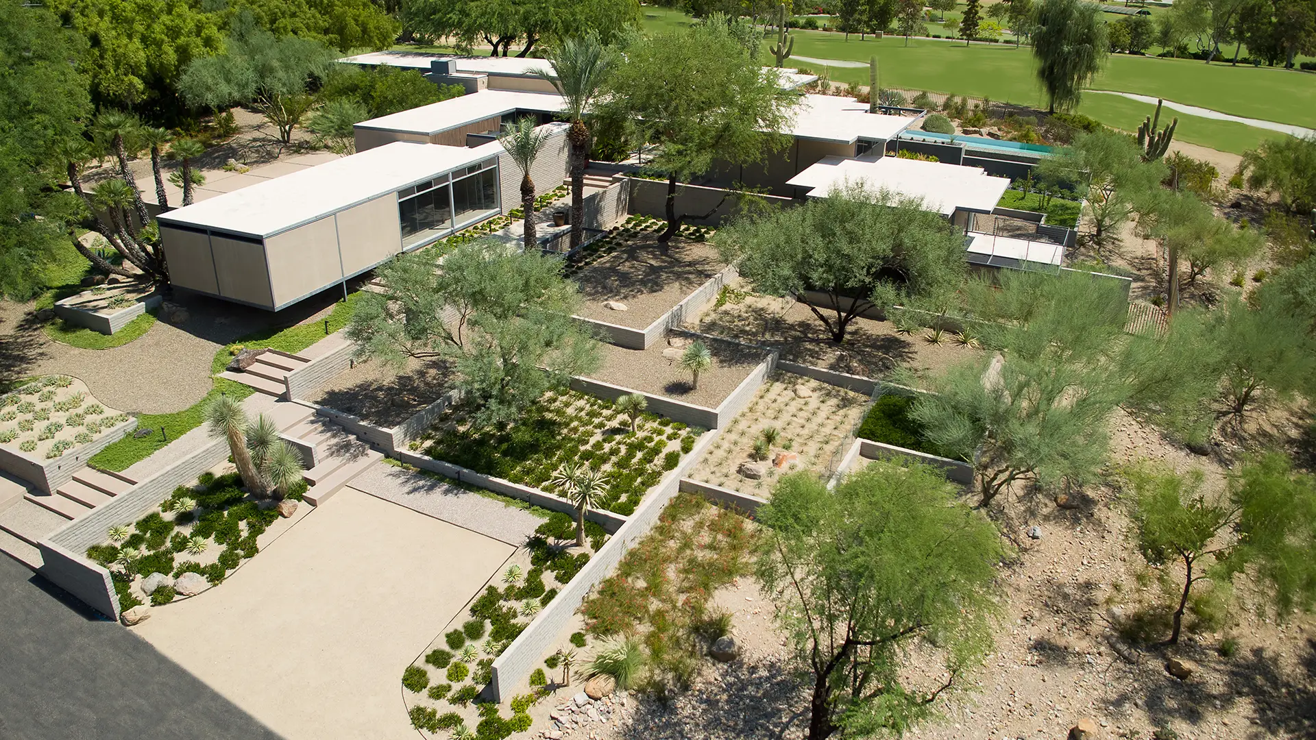 Aerial view of a modern home with tiered landscaping featuring desert plants and trees, surrounded by lush greenery and a golf course.