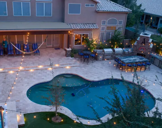 Aerial view of a backyard with a swimming pool, a hot tub, a fire pit, outdoor seating, string lights, a barbecue area, and people standing near lounge chairs.