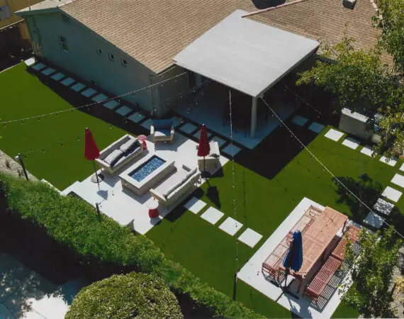 Aerial view of a modern backyard with synthetic turf, a covered patio, a seating area with a fire pit, and a picnic table under an umbrella, surrounded by greenery.