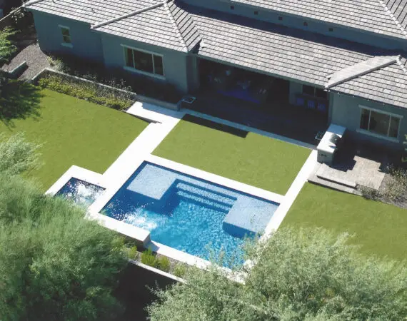 Aerial view of a backyard with a modern rectangular swimming pool and a spa, surrounded by green artificial turf and a paved patio area adjacent to the house.