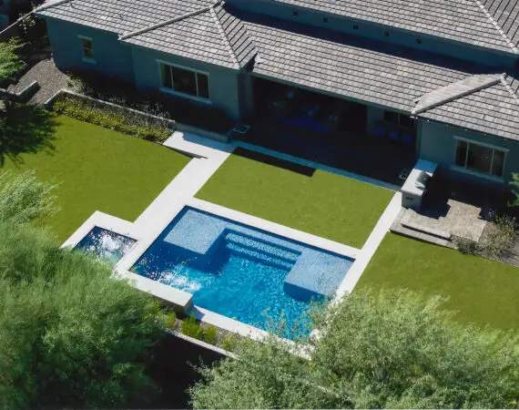 Aerial view of a backyard with a modern rectangular swimming pool and a spa, surrounded by green artificial turf and a paved patio area adjacent to the house.