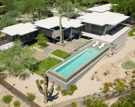 Aerial view of a modern, flat-roofed house with a long, narrow swimming pool, surrounded by desert landscaping with cacti and various greenery.