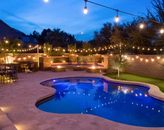 Night view of a backyard with a swimming pool, string lights, outdoor bar with high chairs, and a cozy seating area with fire pits, creating a warm and inviting atmosphere.