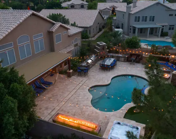 Aerial view of a backyard with a swimming pool, a hot tub, a fire pit, outdoor seating, string lights, a barbecue area, and people standing near lounge chairs.