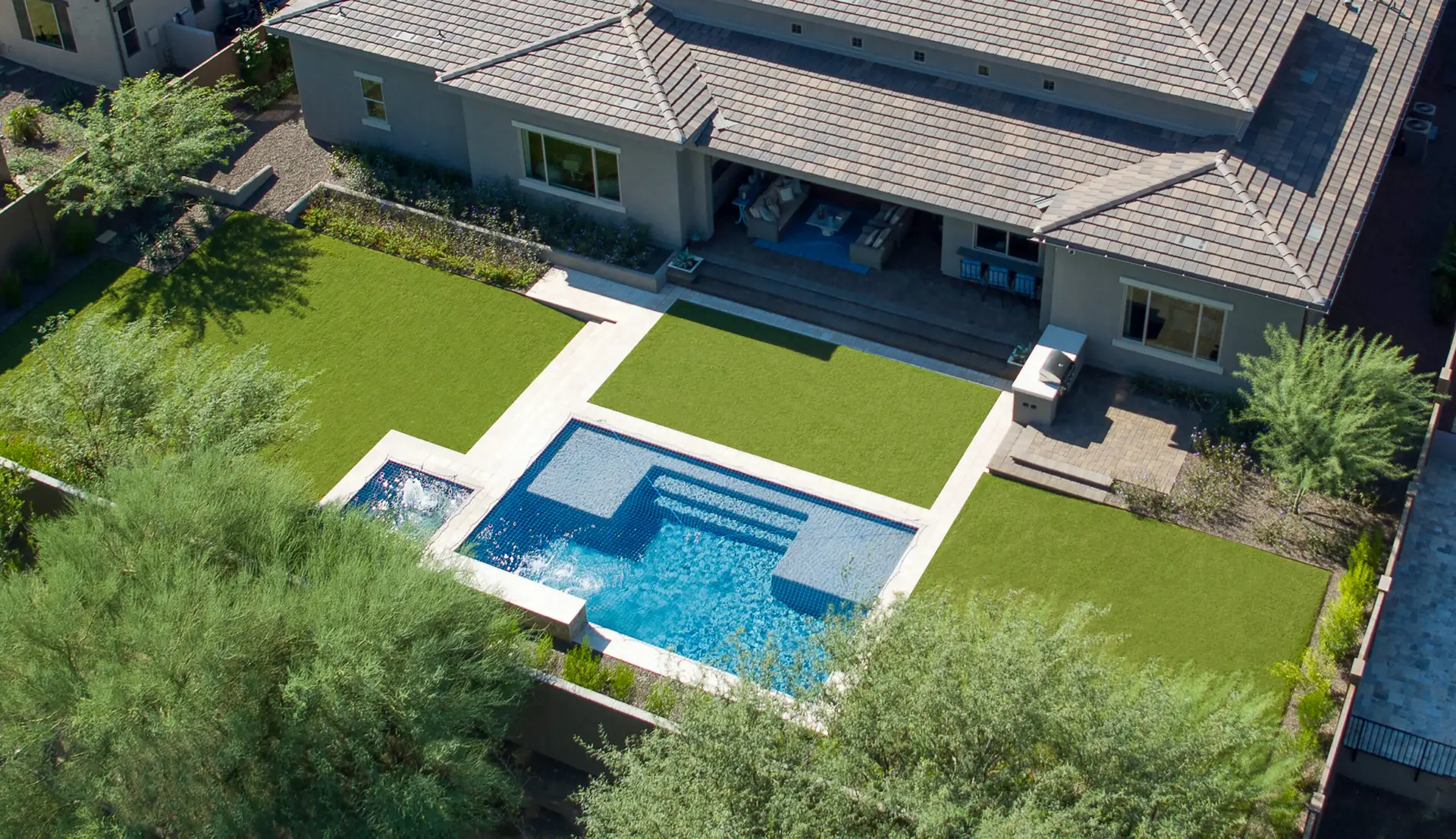 t: Aerial view of a modern backyard with artificial turf, a rectangular pool with a spa, and a covered patio attached to a contemporary house.