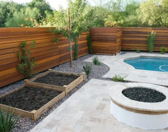 A landscaped backyard with a wooden privacy fence, raised garden beds, a circular fire pit, and a swimming pool with a travertine walkway.