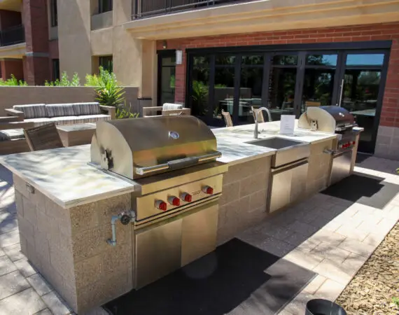 Outdoor barbecue area with stainless steel grills, sinks, and seating, located in a landscaped apartment property.