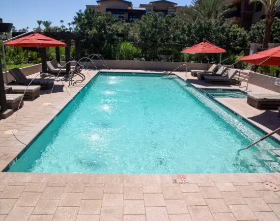 Outdoor pool area with lounge chairs, a red umbrella, and a pergola in the background, surrounded by lush greenery.