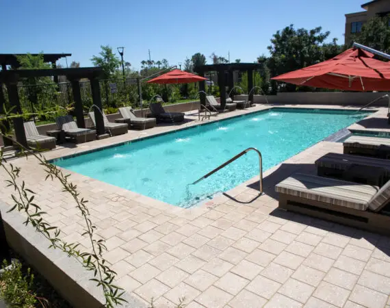 Outdoor pool area with lounge chairs, a red umbrella, and a pergola in the background, surrounded by lush greenery.