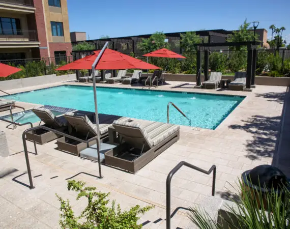 Outdoor pool area with lounge chairs, a red umbrella, and a pergola in the background, surrounded by lush greenery.