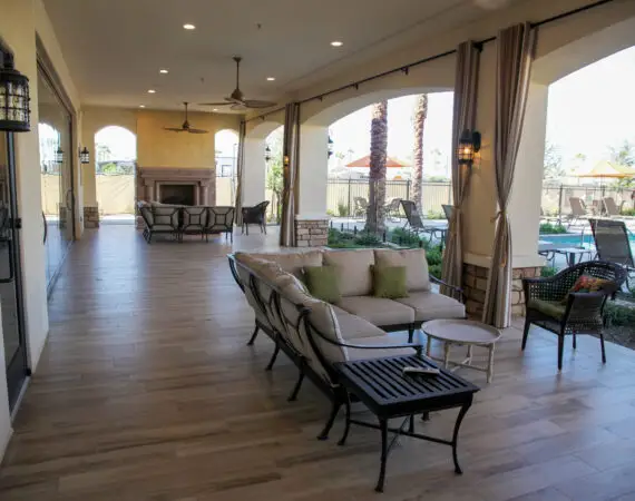 Covered outdoor patio with comfortable seating, ceiling fans, and a fireplace, overlooking a pool area with lounge chairs and umbrellas.