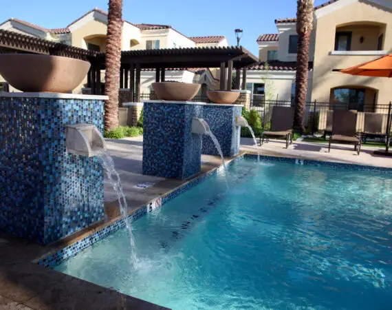 Outdoor pool area with water features and blue mosaic tiles, surrounded by lounge chairs, palm trees, and modern