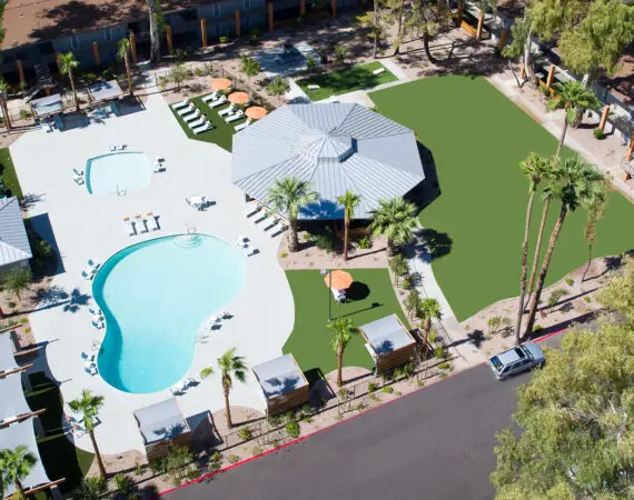 Aerial view of a resort-style swimming pool area with palm trees, lounge chairs, and a pavilion surrounded by landscaped greenery.