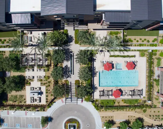 Aerial view of an apartment complex courtyard featuring a swimming pool with red umbrellas, outdoor seating areas, and landscaped walkways surrounded by greenery.