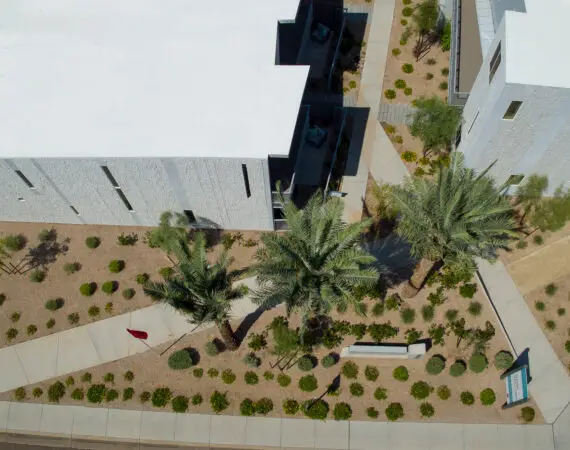 Aerial view of an apartment complex with flat roof, surrounded by landscaped pathways and desert vegetation, including palm trees and various shrubs.