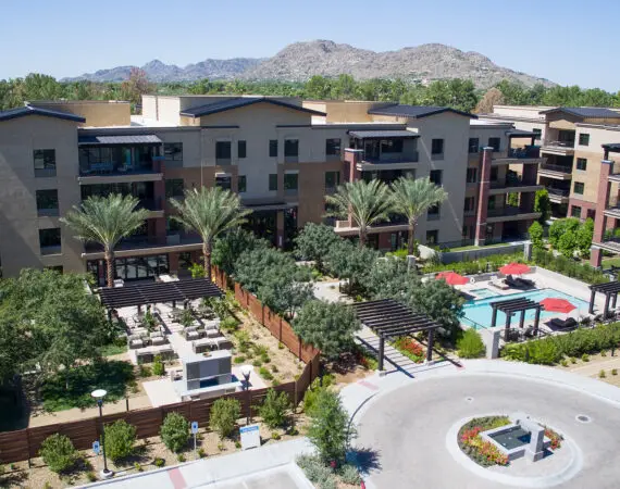 Aerial view of a residential apartment complex with a central swimming pool and surrounding greenery, set against a backdrop of mountainous terrain and a sprawling suburban Phoenix landscape.