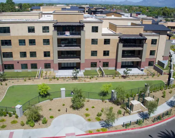 Aerial view of a modern apartment building with a landscaped courtyard and walking paths, surrounded by greenery and set against a suburban backdrop with mountains in the distance.