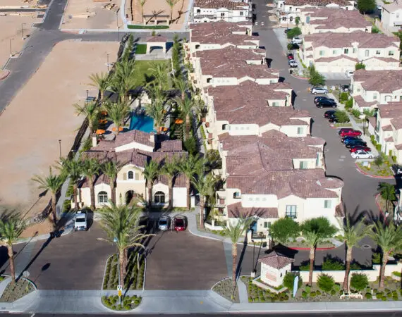 Aerial view of an apartment with tile-roof, a central swimming pool area with palm trees and orange umbrellas, surrounded by a mix of completed and under-construction houses.