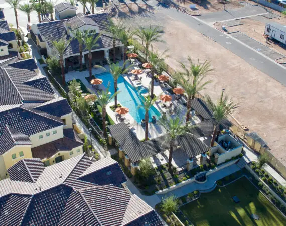 Aerial view of an apartment with tile-roof, a central swimming pool area with palm trees and orange umbrellas, surrounded by a mix of completed and under-construction houses.