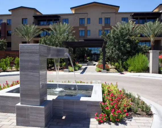 Modern rectangular water fountain in front of a contemporary building, surrounded by vibrant landscaping, trees, and colorful flowers.