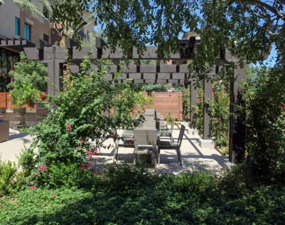 Outdoor dining area with wicker chairs and a long table under a pergola, surrounded by lush green plants and vibrant pink flowers.