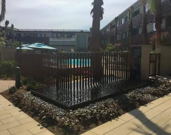 Close-up of a vertical fountain water feature with multiple streams flowing down metal columns, situated in front of a modern building with balconies and windows.