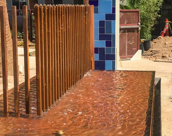 Close-up of a vertical fountain water feature with multiple streams flowing down metal columns, situated in front of a modern building with balconies and windows.