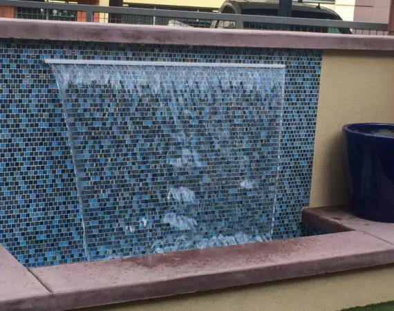 Water feature with cascading water over blue mosaic tiles, bordered by a brown stone ledge and beige wall, with a blue ceramic pot on the side.