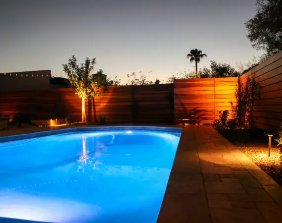 Illuminated backyard pool area at dusk with wooden fencing and subtle garden lighting.