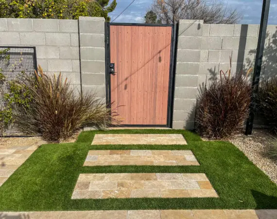 Modern gate with wooden panels framed by black metal, set in a concrete block wall, with a path of stepping stones and green grass leading up to it, flanked by ornamental grasses.