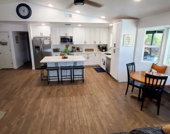 Spacious kitchen with a marble island, white cabinets, wood floors, and a dining nook by large windows. The room includes a water cooler and leads to other areas of the house.