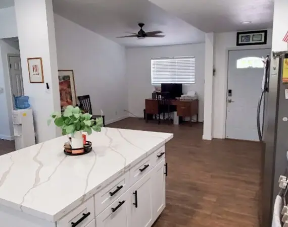 Open kitchen with a marble countertop island, white cabinets, and wood floors. The space leads to a small office area with a desk and chair, and a front door.