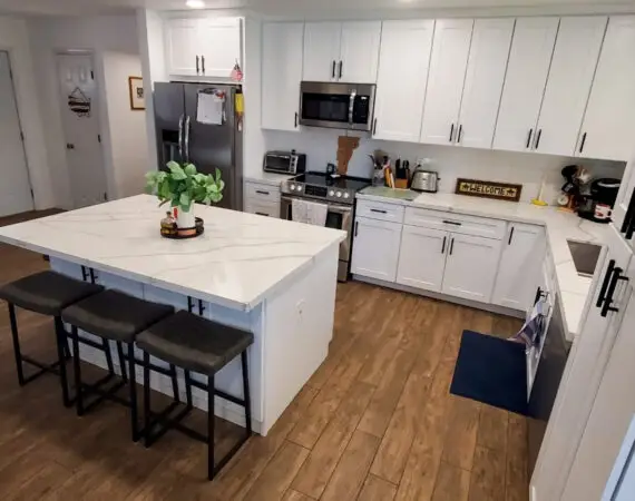 Modern kitchen with a spacious marble island, white cabinets, stainless steel appliances, and wooden flooring, featuring bar stools and a decorative plant.