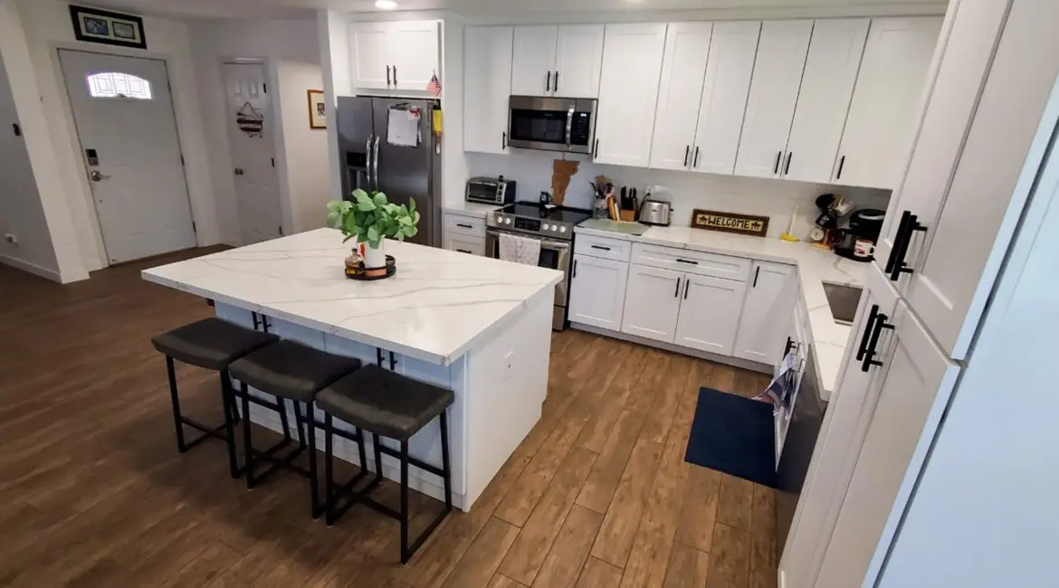 Modern kitchen with a spacious marble island, white cabinets, stainless steel appliances, and wooden flooring, featuring bar stools and a decorative plant.