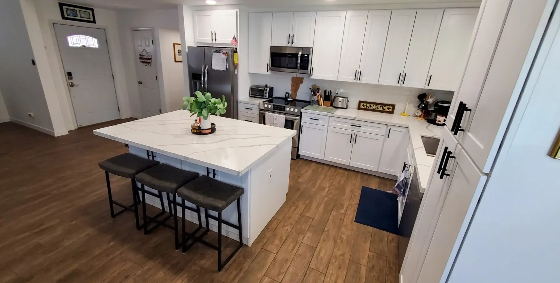 Modern kitchen with a spacious marble island, white cabinets, stainless steel appliances, and wooden flooring, featuring bar stools and a decorative plant.