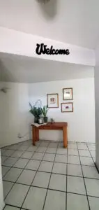 Entryway with white tile floors, a wooden table decorated with plants and framed pictures on the wall. A "Welcome" sign is above the entry.