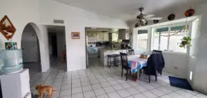 Spacious kitchen and dining area with white tile floors, a table with colorful tablecloth, modern appliances, and large windows allowing natural light, with a small dog present.