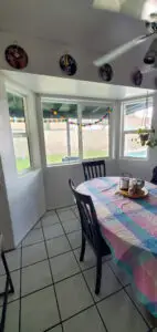 Dining area with a colorful tablecloth, dark wooden chairs, white tile floors, and large windows adorned with a hanging plant and festive decorations.