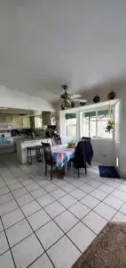 Bright kitchen and dining area with white tile floors, colorful tablecloth on dining table, modern appliances, and large windows bringing in natural light.