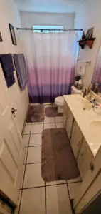 Compact bathroom with a dual-sink vanity, white tile floor, and a shower with a purple gradient curtain. Towels hang on the wall and toiletries are on the counter.