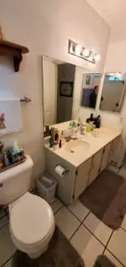 Bathroom with a dual-sink vanity, large mirror, toilet, and white tile floor. The countertop is cluttered with various toiletries and a towel hangs on the wall.