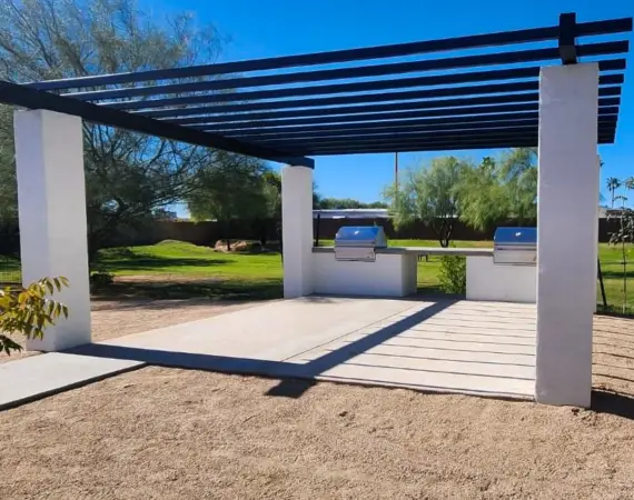 Modern outdoor grill area with a sleek pergola, dual built-in grills, and a spacious concrete patio set in a serene park-like setting.