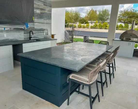 Modern outdoor kitchen with large marble countertop island, wicker bar stools, built-in stainless steel oven, sink, and white cabinetry overlooking a landscaped backyard.